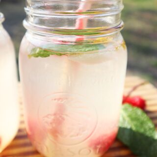 Mason jar with raspberry mint lemonade with dandelions sitting on a bamboo cutting board and cut fruit laying around it. The cup has a pink and white striped straw.