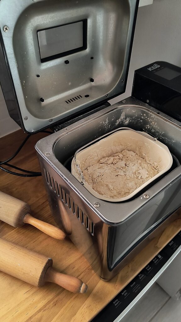 Bread machine with pizza dough mixing and two wooden rolling pins beside the machine.