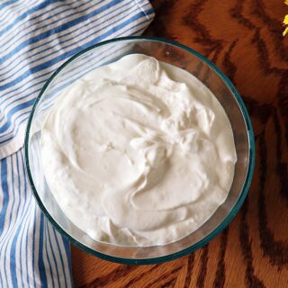 Overhead shot of a bowl of instant pot greek yogurt on a wood table with kitchen towel to the left and a vase of wildflowers to the right