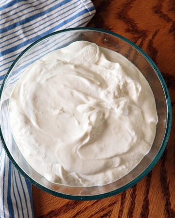 Overhead shot of a bowl of instant pot greek yogurt on a wood table with kitchen towel to the left and a vase of wildflowers to the right