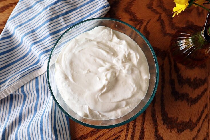 Overhead shot of a bowl of instant pot greek yogurt on a wood table with kitchen towel to the left and a vase of wildflowers to the right