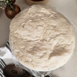 Bowl of pizza dough rising with a vase of wildflowers beside it and a kitchen towel with wood spoon on the other side.