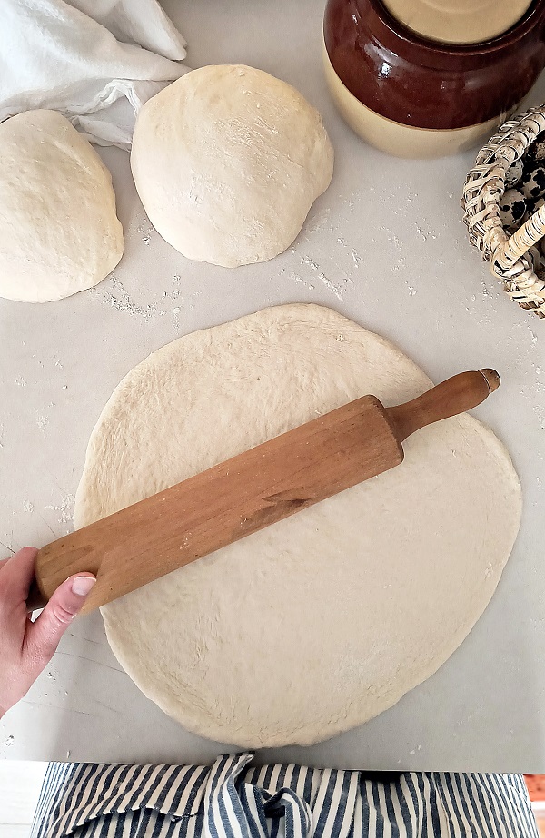 woman rolling out pizza dough on countertop with balls of pizza dough to the side.