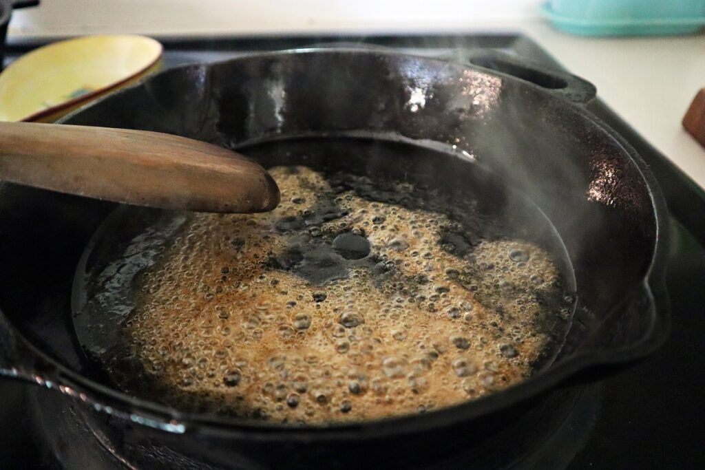 Wood spoon stirring maple syrup and avocado oil together in an iron skillet for quick skillet granola. The mixture is hot and bubbly.