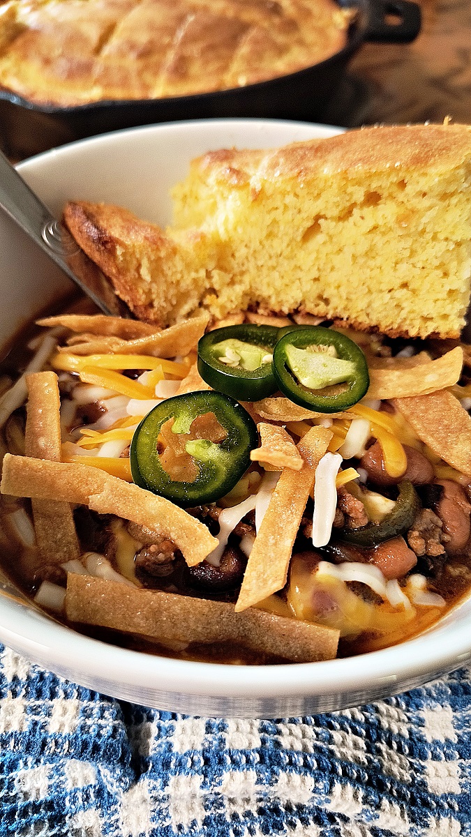 Bowl of homemade chili with toppings and cornbread sitting on a blue and white checkered kitchen towel.