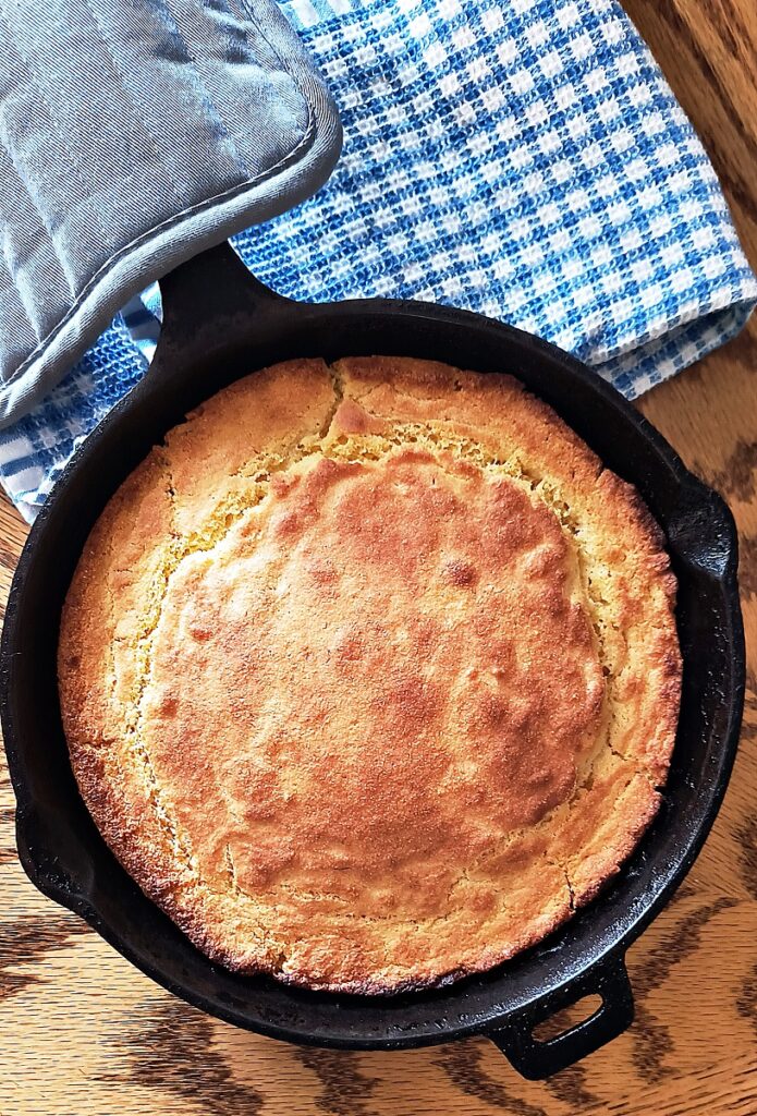 Iron skillet with crispy cornbread. An oven mitt is laying across the handle and there is a checkered blue and white towel underneath.