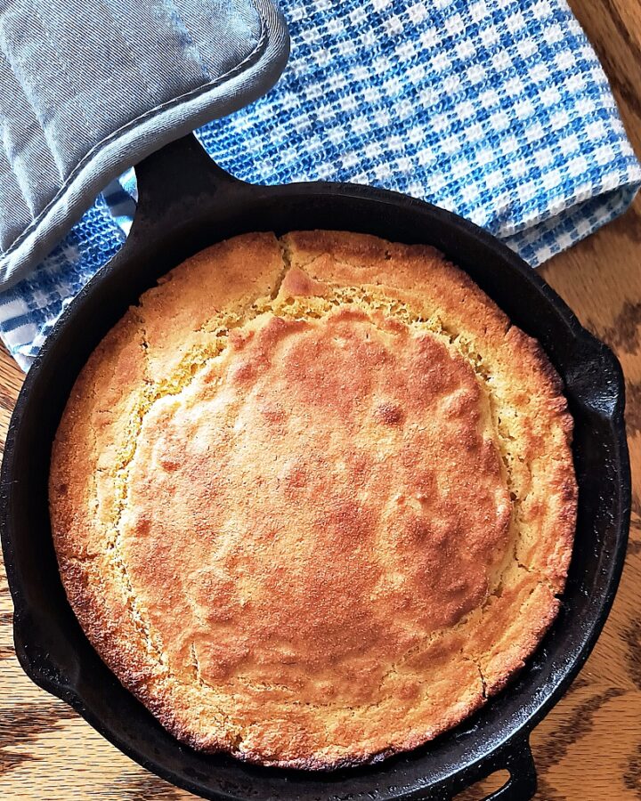 Overhead shot of an iron skillet with cornbread and a blue checkered towel beside it.