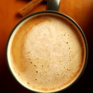 Top view of coffee mug with snickerdoodle coffee creamer foam on top and cinnamon sticks in the background on wood counter surface.