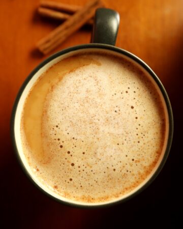 Top view of coffee mug with snickerdoodle coffee creamer foam on top and cinnamon sticks in the background on wood counter surface.