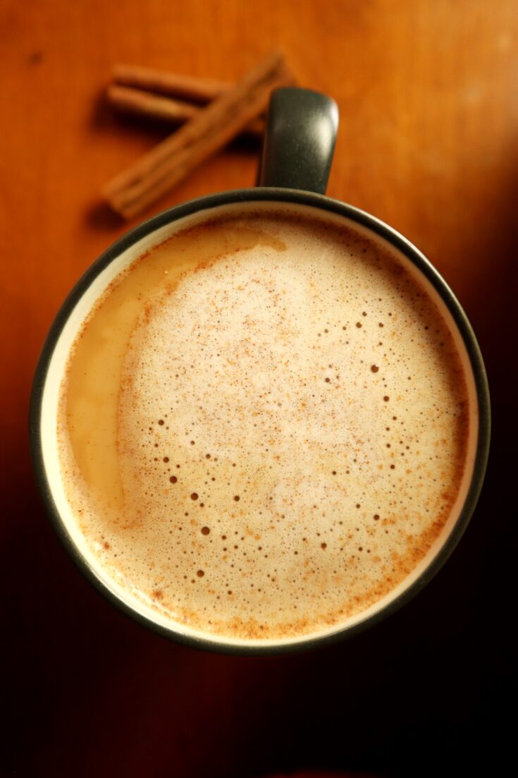 Top view of coffee mug with snickerdoodle coffee creamer foam on top and cinnamon sticks in the background on wood counter surface.