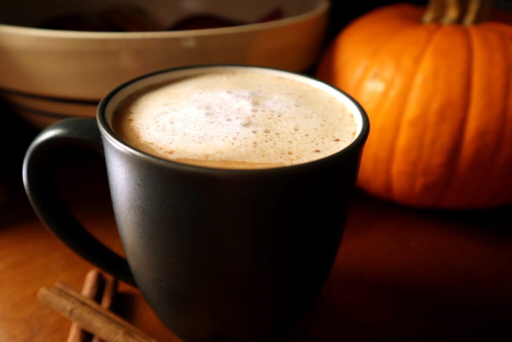 Coffee mug with snickerdoodle coffee creamer foam on top with cinnamon sticks beside the mug and a pumpkin in the background.